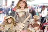 Several vintage dolls are displayed prominently, with the central doll wearing a detailed outfit including a dress, apron, and hat. Surrounding it are other dolls, each with distinct hairstyles and clothing, creating a nostalgic and charming scene. The background suggests an outdoor market setting with people blurred out.