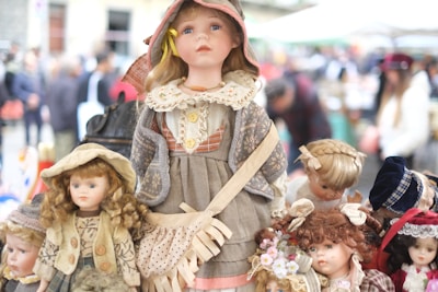 Several vintage dolls are displayed prominently, with the central doll wearing a detailed outfit including a dress, apron, and hat. Surrounding it are other dolls, each with distinct hairstyles and clothing, creating a nostalgic and charming scene. The background suggests an outdoor market setting with people blurred out.