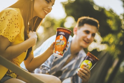 Two people enjoying drinks outdoors, holding cups labeled 'Michelista' with vibrant designs. The scene is warmly lit, highlighting their casual and relaxed expressions. The background is a soft blur of greenery, suggesting a park or garden setting.