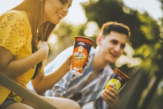Two people enjoying drinks outdoors, holding cups labeled 'Michelista' with vibrant designs. The scene is warmly lit, highlighting their casual and relaxed expressions. The background is a soft blur of greenery, suggesting a park or garden setting.