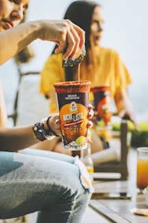 A person is pouring a packet into a Michelada mix cup with a red rim, while another person in the background sits wearing a yellow shirt. The ambiance is casual and likely outdoors.