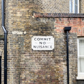 A vintage brick wall features a white sign with bold black letters saying 'COMMIT NO NUISANCE.' The wall is composed of aged bricks in shades of yellow, brown, and red, with a black drainpipe to the left and a window to the right, partially visible with white framing. The atmosphere reflects a nostalgic urban setting.
