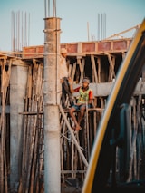 Two construction workers are engaged in work at a building site. One worker is standing on wooden scaffolding, while the other is seated on the scaffolding structure. They appear to be communicating or working together on the task at hand. The construction site is surrounded by wooden planks and metal rods, indicating an unfinished building structure.