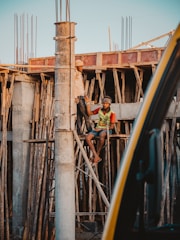 Two construction workers are engaged in work at a building site. One worker is standing on wooden scaffolding, while the other is seated on the scaffolding structure. They appear to be communicating or working together on the task at hand. The construction site is surrounded by wooden planks and metal rods, indicating an unfinished building structure.