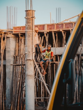Two construction workers are engaged in work at a building site. One worker is standing on wooden scaffolding, while the other is seated on the scaffolding structure. They appear to be communicating or working together on the task at hand. The construction site is surrounded by wooden planks and metal rods, indicating an unfinished building structure.