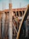 Two construction workers are engaged in work at a building site. One worker is standing on wooden scaffolding, while the other is seated on the scaffolding structure. They appear to be communicating or working together on the task at hand. The construction site is surrounded by wooden planks and metal rods, indicating an unfinished building structure.