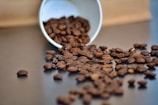 Coffee beans are scattered across a smooth surface, spilling out from a tipped-over white cup. The beans have a rich, dark brown color and create a textured pattern against the neutral background.