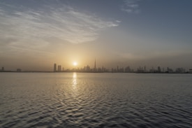 A serene sunrise over a city skyline with tall skyscrapers silhouetted against the light. The sun is partially hidden by the buildings, casting a warm glow across the sky and reflecting on the water in the foreground.