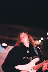 A person with long hair playing an electric guitar on stage, wearing a black hoodie with 'INFLUENCE' written on it. The background is dark with stage lights creating a vibrant atmosphere.