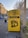 A yellow donation bin for clothes and shoes is situated on a city sidewalk, covered with graffiti. The bin is labeled with the names of several organizations, along with instructions for use. Surrounding the bin are some buildings, a road with vehicles, a streetlight, and a metal fence.