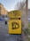 A yellow donation bin for clothes and shoes is situated on a city sidewalk, covered with graffiti. The bin is labeled with the names of several organizations, along with instructions for use. Surrounding the bin are some buildings, a road with vehicles, a streetlight, and a metal fence.
