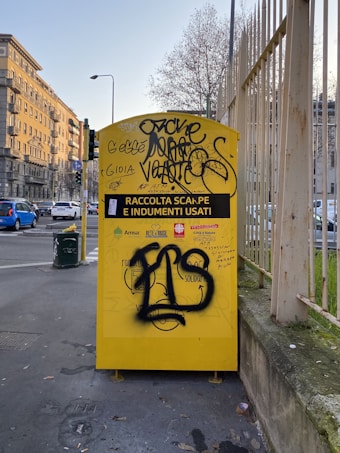 A yellow donation bin for clothes and shoes is situated on a city sidewalk, covered with graffiti. The bin is labeled with the names of several organizations, along with instructions for use. Surrounding the bin are some buildings, a road with vehicles, a streetlight, and a metal fence.