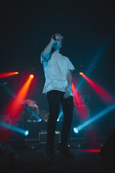 A performer is on stage holding a microphone, dressed in a white t-shirt and dark pants. The stage is lit with vibrant red and blue spotlights creating a dramatic and energetic atmosphere.