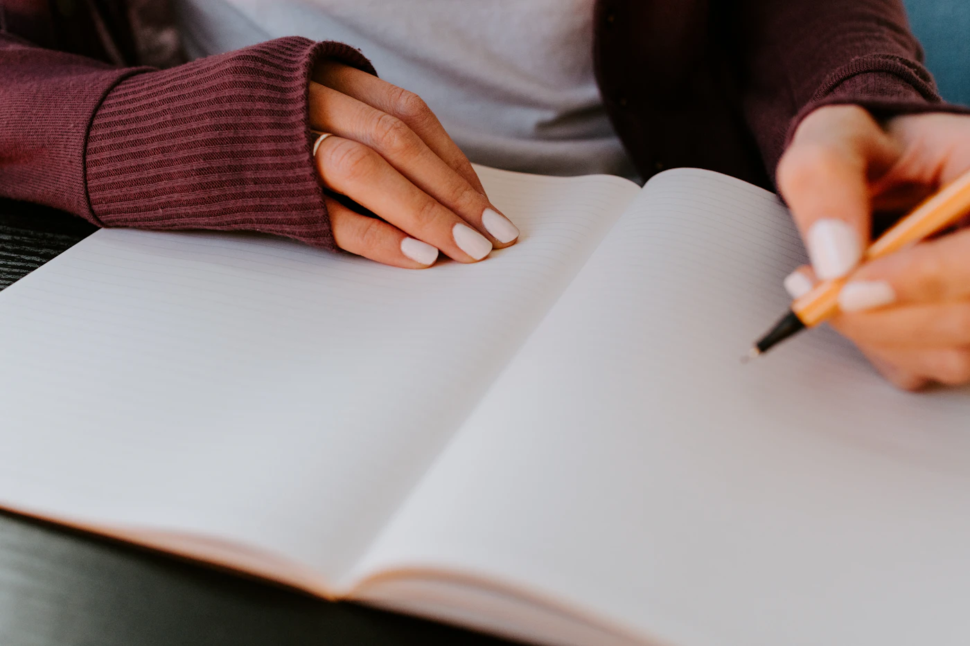 Mãos de uma mulher de cardigã vinho escrevendo em um caderno aberto de páginas em branco