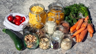 A collection of fresh vegetables and dry goods displayed on a textured surface. The image includes a variety of glass jars filled with nuts, seeds, and pasta. There are whole carrots with green tops, a zucchini, and a bag of ripe red tomatoes. The jars are arranged in a group, showcasing their contents.