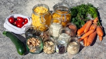 A collection of fresh vegetables and dry goods displayed on a textured surface. The image includes a variety of glass jars filled with nuts, seeds, and pasta. There are whole carrots with green tops, a zucchini, and a bag of ripe red tomatoes. The jars are arranged in a group, showcasing their contents.