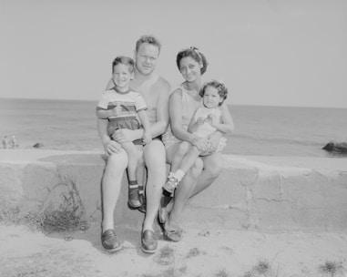 A family of four sits together on a stone wall overlooking the ocean. The father and mother sit in the center, with the father holding a young boy on his knee and the mother holding a young girl. The beach setting suggests it is a sunny day, and the family appears to be enjoying a leisurely outing.