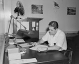 A man is seated at a desk in an office environment, concentrating on writing on a piece of paper. The desk has several items, including a lamp, some paperwork, an ashtray, and a fan. The walls are decorated with photographs of landscapes and a small plant ornament.