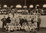 A group of early 20th-century baseball players poses for a photograph on a field. The team members are wearing uniforms with different letters and names, such as Michigan and ACA. They are surrounded by a large crowd of spectators in the background, seated in a grandstand, dressed in period-appropriate formal attire, including hats and suits.