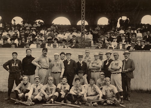 A group of early 20th-century baseball players poses for a photograph on a field. The team members are wearing uniforms with different letters and names, such as Michigan and ACA. They are surrounded by a large crowd of spectators in the background, seated in a grandstand, dressed in period-appropriate formal attire, including hats and suits.
