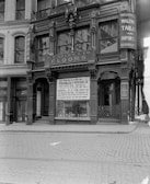 A black and white photograph of a historic building with ornate architectural details, displaying signage for R.T. Adams & Co. Floors and Walter Tailor and Importer. The storefront features large windows with promotional posters for a footwear company, Franklin P. Winston Co., advertising a removal sale. Cobblestone streets are visible in front of the building.