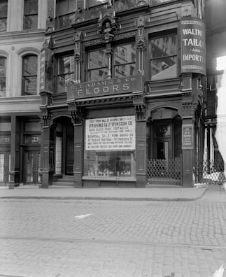 A black and white photograph of a historic building with ornate architectural details, displaying signage for R.T. Adams & Co. Floors and Walter Tailor and Importer. The storefront features large windows with promotional posters for a footwear company, Franklin P. Winston Co., advertising a removal sale. Cobblestone streets are visible in front of the building.