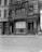 A black and white photograph of a historic building with ornate architectural details, displaying signage for R.T. Adams & Co. Floors and Walter Tailor and Importer. The storefront features large windows with promotional posters for a footwear company, Franklin P. Winston Co., advertising a removal sale. Cobblestone streets are visible in front of the building.
