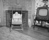 A baby sits joyfully in a stroller in front of a brick fireplace. To the right, an old-fashioned television set is placed on a small table next to a pair of decorative fireplace tools. A patterned rug covers part of the wooden floor.