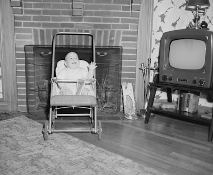 A baby sits joyfully in a stroller in front of a brick fireplace. To the right, an old-fashioned television set is placed on a small table next to a pair of decorative fireplace tools. A patterned rug covers part of the wooden floor.