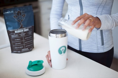A person is pouring liquid from a glass bottle into a white shaker or tumbler with a green emblem on the side. A protein or recovery shake package labeled 'Lyfefuel' is nearby on the countertop. The person is wearing a light gray zip-up athletic top, and a separate shaker lid rests on the surface.