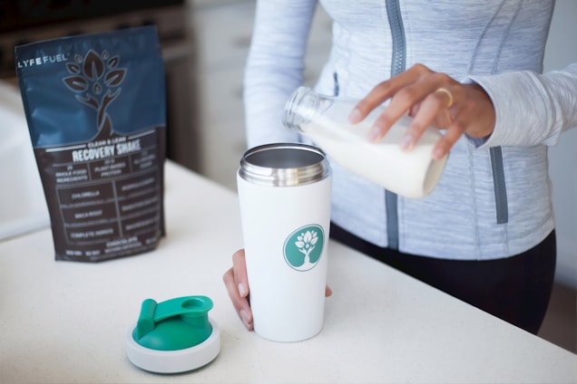 A person is pouring liquid from a glass bottle into a white shaker or tumbler with a green emblem on the side. A protein or recovery shake package labeled 'Lyfefuel' is nearby on the countertop. The person is wearing a light gray zip-up athletic top, and a separate shaker lid rests on the surface.