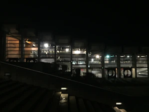 A dimly lit building with large windows revealing an interior illuminated by bright artificial lights. The structure features columns and appears to be part of a modern architectural design. In the foreground, there is a staircase with lights embedded within the steps.