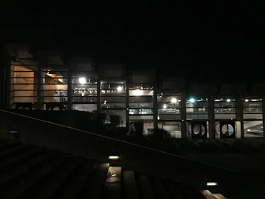 A dimly lit building with large windows revealing an interior illuminated by bright artificial lights. The structure features columns and appears to be part of a modern architectural design. In the foreground, there is a staircase with lights embedded within the steps.