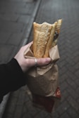 A hand holding a paper bag containing a baguette, with part of the bread exposed. The background consists of a paved street with a muted color palette, suggesting an outdoor setting. The hand is wearing a silver ring and a black sleeve.