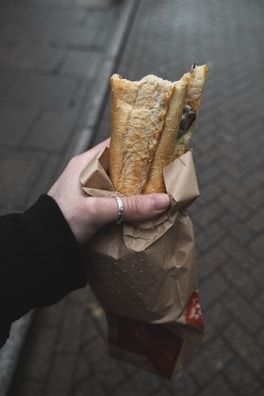 A hand holding a paper bag containing a baguette, with part of the bread exposed. The background consists of a paved street with a muted color palette, suggesting an outdoor setting. The hand is wearing a silver ring and a black sleeve.