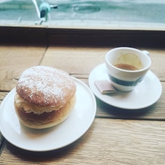 A wooden table near a window displays a dessert pastry topped with powdered sugar on a white plate, accompanied by a small cup of espresso on a matching saucer with a small piece of chocolate.
