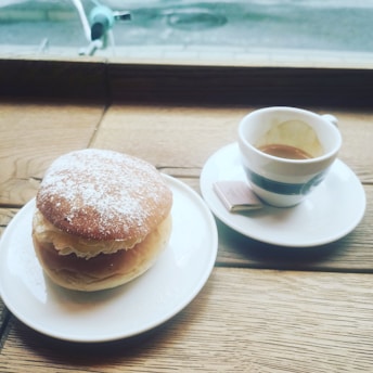A wooden table near a window displays a dessert pastry topped with powdered sugar on a white plate, accompanied by a small cup of espresso on a matching saucer with a small piece of chocolate.
