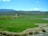 A vast expanse of green fields extends into the distance, with a small herd of horses grazing. A solitary red barn with a white roof is situated near the center, surrounded by rolling hills under a bright blue sky with a few clouds.