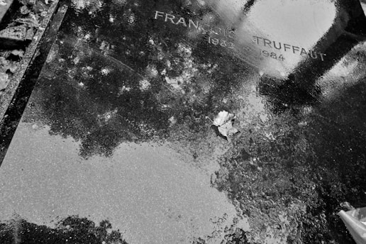A black and white photograph of a wet grave surface with the name and dates indicating it belongs to someone who lived from 1932 to 1984. One dry leaf is visible on the glossy, reflective surface that also shows the reflection of trees above.