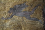 A mosaic artwork featuring a dark, winged figure in motion against a backdrop of small, multicolored tiles. The figure appears to be mythical, with elongated limbs and horns, conveying a sense of dynamic movement.