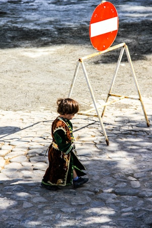 A young child in elaborate, traditional clothing walks on a cobblestone path. The child is adorned in a velvet costume with intricate details and colorful beads. Nearby, a red and white traffic sign stands on a metal frame, casting a shadow on the ground.