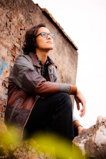 A person with glasses and long hair looks upwards contemplatively while sitting against a rustic, textured wall. Dressed in a plaid shirt and a brown and gray jacket, the individual is surrounded by rock formations in an outdoor setting.