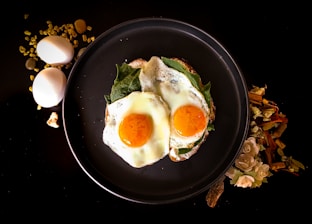 Two sunny-side-up eggs are placed on a bed of greens on a dark round plate. To the left of the plate, two whole eggs and some golden granules are scattered on a black surface. To the right, there is a decorative arrangement of dried flowers and wooden sticks.
