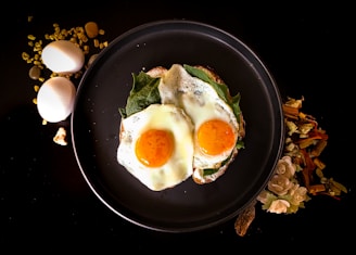 Two sunny-side-up eggs are placed on a bed of greens on a dark round plate. To the left of the plate, two whole eggs and some golden granules are scattered on a black surface. To the right, there is a decorative arrangement of dried flowers and wooden sticks.