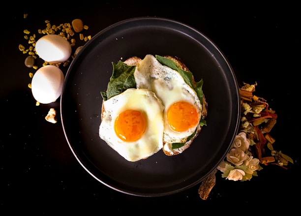 Two sunny-side-up eggs are placed on a bed of greens on a dark round plate. To the left of the plate, two whole eggs and some golden granules are scattered on a black surface. To the right, there is a decorative arrangement of dried flowers and wooden sticks.