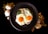 Two sunny-side-up eggs are placed on a bed of greens on a dark round plate. To the left of the plate, two whole eggs and some golden granules are scattered on a black surface. To the right, there is a decorative arrangement of dried flowers and wooden sticks.