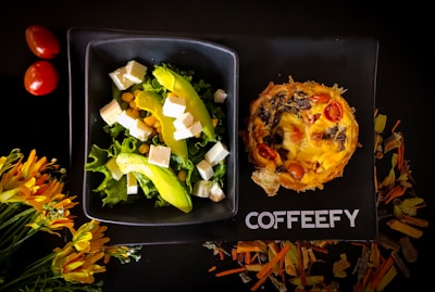 A stylish black plate holds a vibrant salad with lettuce, avocado slices, cubes of white cheese, and corn, alongside a savory pie topped with tomatoes and mushrooms. Nearby, two red cherry tomatoes and a cluster of yellow-orange flowers complement the setup. The word 'COFFEEFY' is printed on the plate.
