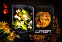 A stylish black plate holds a vibrant salad with lettuce, avocado slices, cubes of white cheese, and corn, alongside a savory pie topped with tomatoes and mushrooms. Nearby, two red cherry tomatoes and a cluster of yellow-orange flowers complement the setup. The word 'COFFEEFY' is printed on the plate.