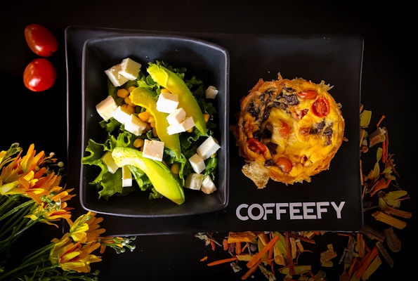 A stylish black plate holds a vibrant salad with lettuce, avocado slices, cubes of white cheese, and corn, alongside a savory pie topped with tomatoes and mushrooms. Nearby, two red cherry tomatoes and a cluster of yellow-orange flowers complement the setup. The word 'COFFEEFY' is printed on the plate.