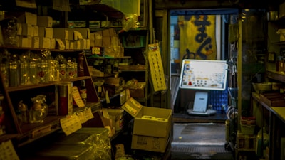 A dimly lit, narrow aisle in a traditional store, with shelves lined with various goods including canned items, packaging materials, and boxes. The entrance at the far end of the aisle opens to a small area outside, with a large yellow sign displaying some Japanese characters and a bright vending machine, suggesting a busy, industrious atmosphere.
